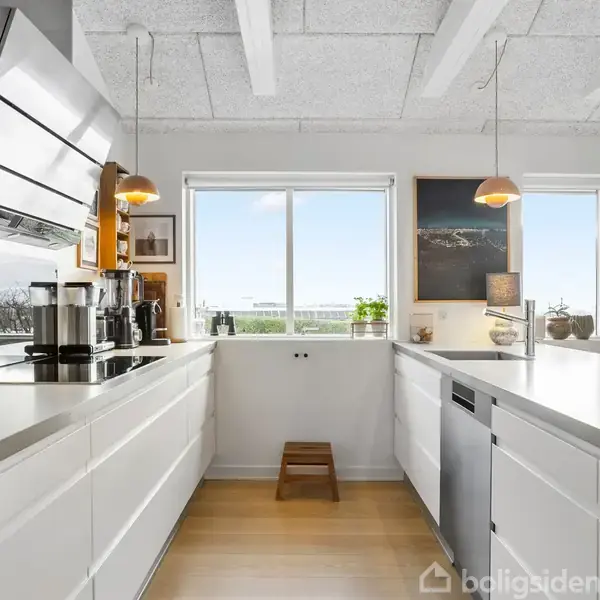 Et lyst køkken med hvide skabe og trægulv. Køkkenet har moderne apparater, en emhætte, og store vinduer der lukker naturligt lys ind. Små planter pryder vindueskarmen.