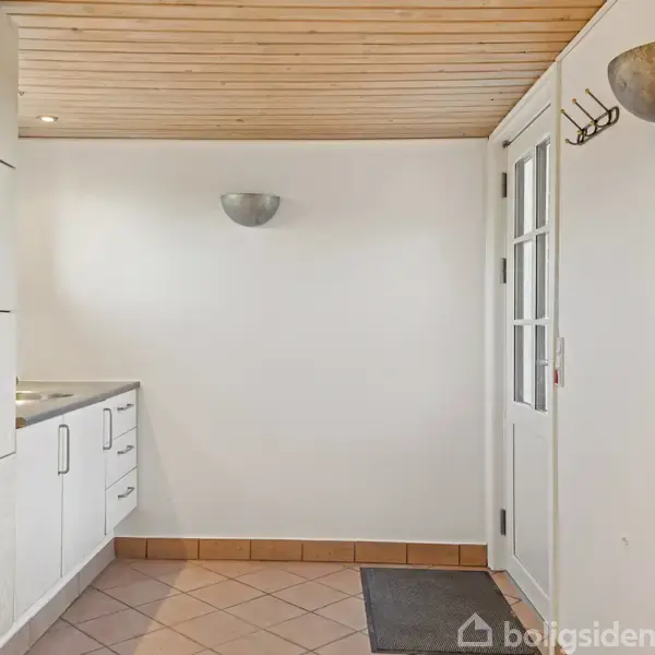 Vaskemaskine og skabe står mod venstre væg i et rum med flisegulv og træloft. Hvide vægge har en dør og nogle knager til højre.