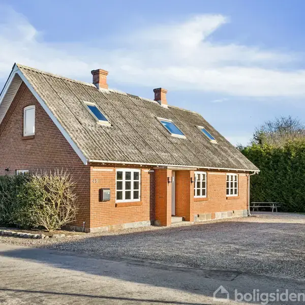 Hus af røde mursten med skråt tag og kviste, står stille på en gruset vej omgivet af hække og blå himmel.