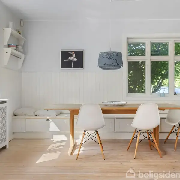 Spisebord med fire hvide stole i et lyst rum. Et stort vindue giver udsigt til grønne omgivelser. Hylde med bøger og billede af ballerina på væggen.