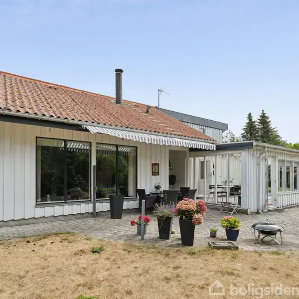 Et hvidt, moderne hus med rødt tegltag står i en have med stenet terrasse. Blomsterkrukker og havestole er placeret foran, omgivet af græs og høje træer.