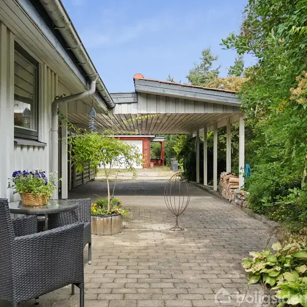 Et hvidt hus med rødt tag står på en stor græsplæne. Solstole er placeret i haven omgivet af træer og buske under en blå himmel.