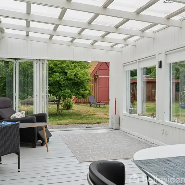 En overdækket terrasse med transparente vægge indeholder loungemøbler. Udenfor ses en grøn have med et træ og røde træbygninger. En fuglebure hænger i hjørnet.