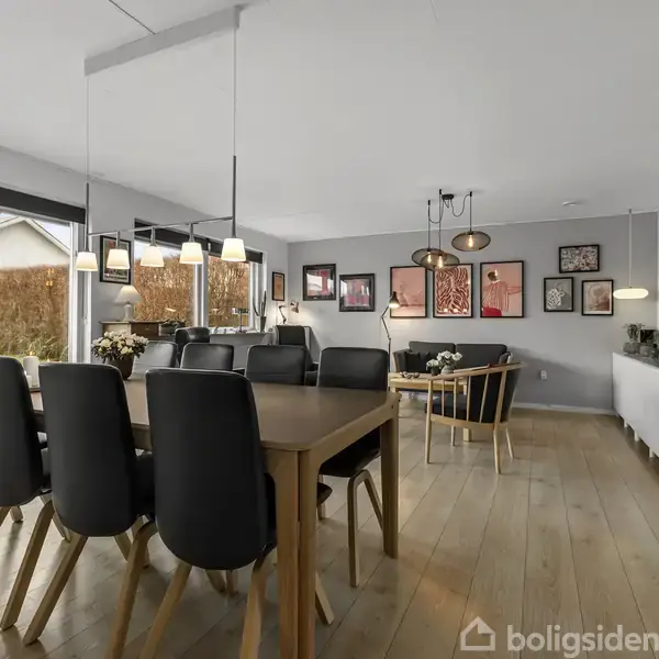 En stor spisestue med et langt træbord omgivet af sorte stole. Store vinduer slipper lys ind, og der er kunst på væggene. Skabe står langs højrevæggen.