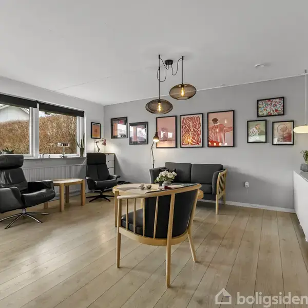 Stue med grå sofa og to sorte lænestole står på trægulv. Væggene er dekoreret med farverige billeder. Et stort vindue til venstre giver naturligt lys. Indretningsdetaljer inkluderer en hvid skænk og lamper.