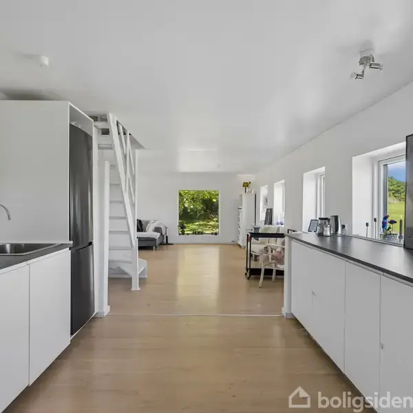 Køkkenet med hvide skabe og sort bordplade strækker sig langs væggen. Trappe i baggrunden fører til øvre etage. Store vinduer giver udsigt til grønne omgivelser.