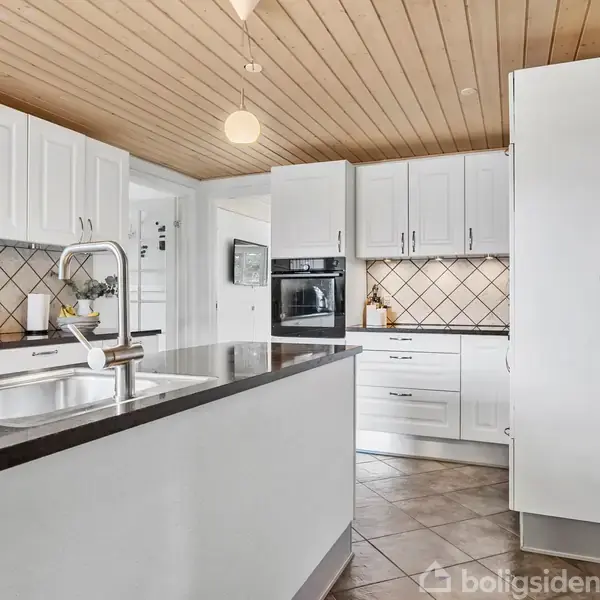 Et moderne køkken med hvide skabe udført i træ, et mørkt bordplade med integreret vask, omgivet af flisebelagt væg og hvidevarer. Lysebrunt loft med nedhængt lampe.