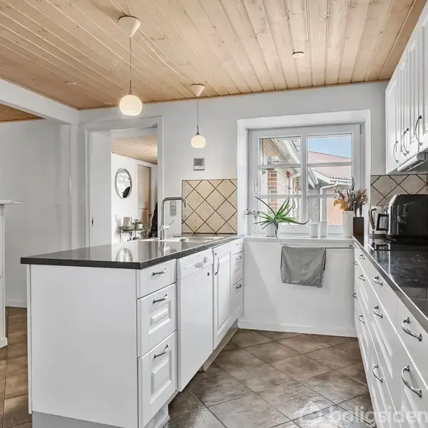 Rødstenshus med skråt tag og tilhørende garage står i en gårdsplads. Blå himmel og græsplæne omkranser ejendommen.