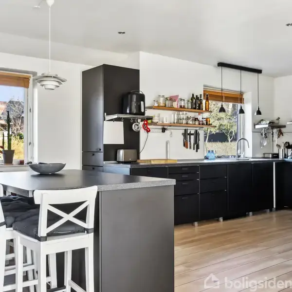 Sort køkkenbord med hvide stole står i køkkenområde med sorte skabe og hylder. Store vinduer bringer naturligt lys ind, planter pynter bordet, og der ses køkkenredskaber og apparater.