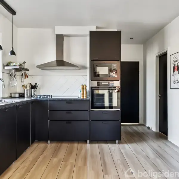 Sort køkken med indbyggede ovne og emhætte, lyse trægulve og stål-køleskab. Hængende lamper over vasken. Tegninger på væggen. Lyse vinduer giver udsigt til haven.