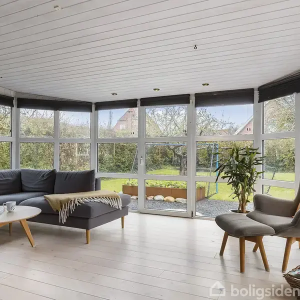 En stue med sofa og lænestol står vendt mod store vinduer, der giver udsigt til en have. Runde sofabord og planter giver en hyggelig atmosfære.