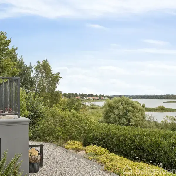 En balkon med sorte møbler står til venstre. Det grusbelagte område foran fører til buske og træer. I baggrunden ses en bred flod under en klar himmel.