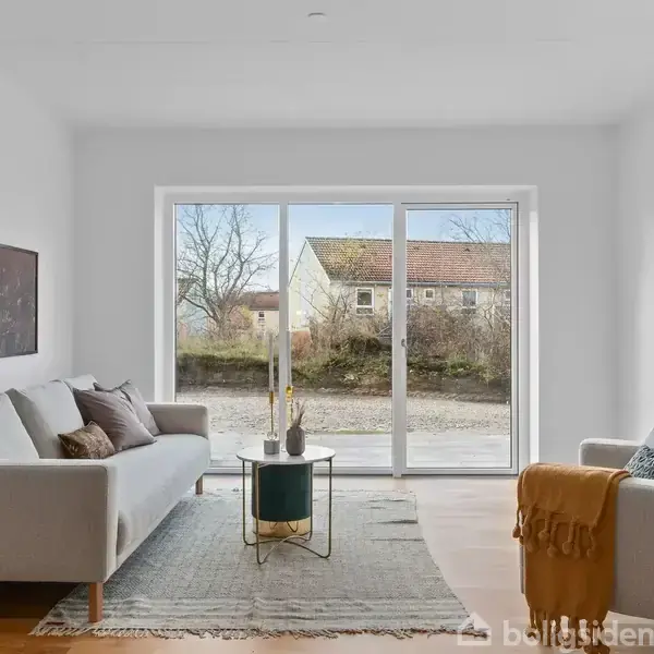 Stue med to sofaer, et lavt sofabord og en lampe, der står ved et stort vindue med udsigt til en gård og et hus i baggrunden.