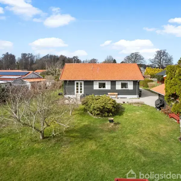 Hus med rødt tegltag står på grøn græsplæne med træer, omgivet af andre huse i et villakvarter under blå himmel med skyer.