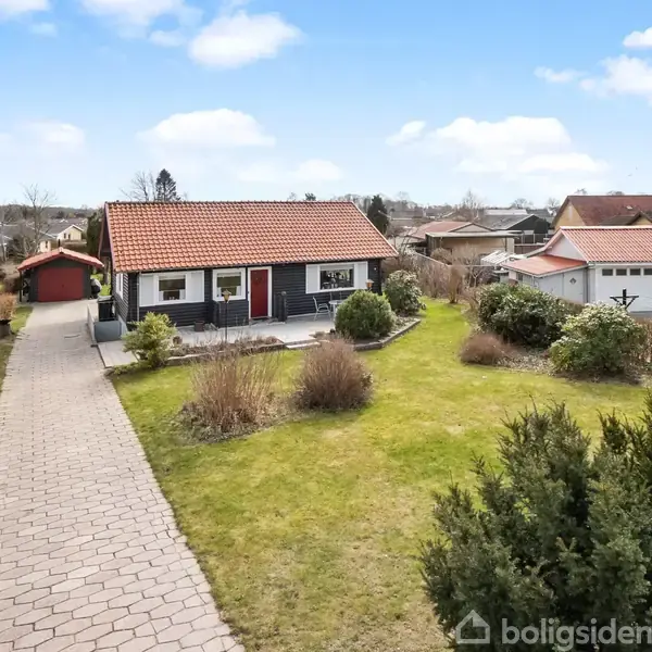 Et lille hus med rødt tag står omgivet af en velplejet have og stenbelagt sti. Ved siden af er der en rød carport. Skyfri blå himmel i baggrunden.