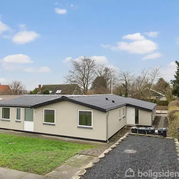 En lav, beige enetages hus med fladt tag står på en velplejet græsplæne. En grussti fører til indgangen. Buske og træer omgiver ejendommen, og himlen er let skyet.