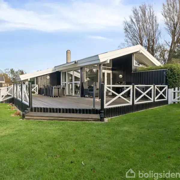 Et sort træhus med hvide detaljer har en stor træterrasse. Terrassen er omgivet af et grønt græsareal, mens høje træer er synlige i baggrunden.