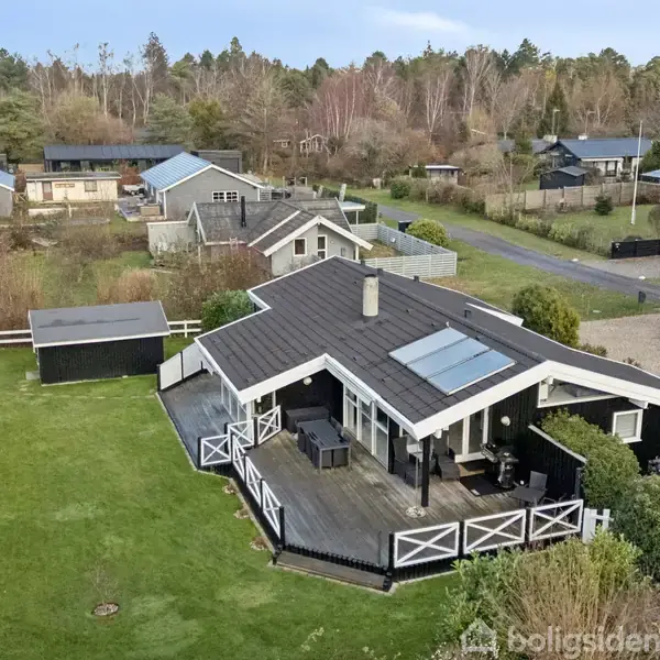 Sommerhus med sort tag og veranda står i en grøn have omkranset af et hegn. Flere træer og andre huse ses i baggrunden.