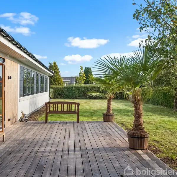 En træterrasse med en træbænk og to palmer i krukker står foran et hus. Baggrunden viser en græsplæne og en hæk under en blå himmel med skyer.