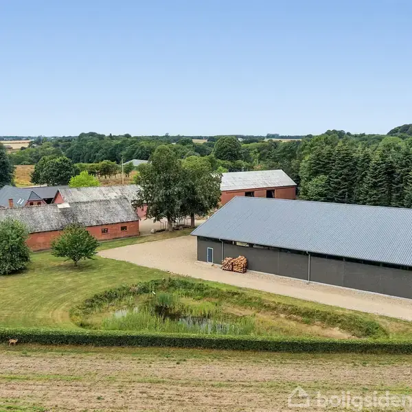 En gård med flere bygninger står stille omgivet af åbne marker og skov. Foran ligger en dam omgivet af grønne plæner og enkelte træer.