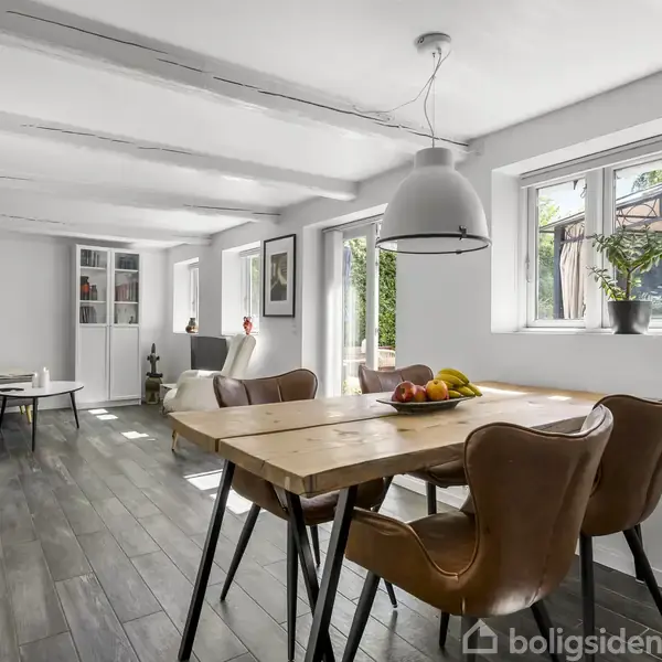 Et moderne spisestue med et træbord og læderstole. En stor lampe hænger over bordet. Væggene er hvide med store vinduer, der slipper naturligt lys ind. En frugtskål står på bordet.