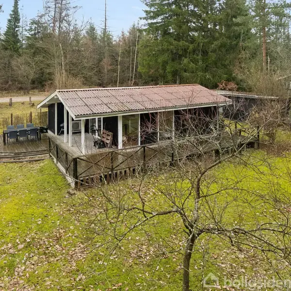Et træhus står stille i en skovklædt have med grønt græs og spredte træer. En terrasse med havemøbler omringer huset, og skove omkranser grunden.
