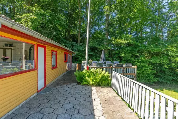 En gul træbygning med røde dørkarme står ved siden af en terrasse. Terrassen har borde og stole og er omgivet af frodige grønne træer.