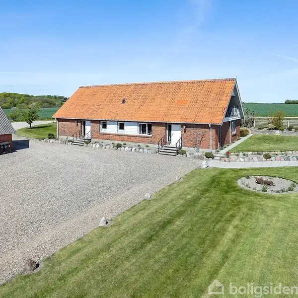 En murstensbygning med rødt tag står i et landligt område. Bygningen er omgivet af en grusplads og velplejet græsplæne med en rund bed.
