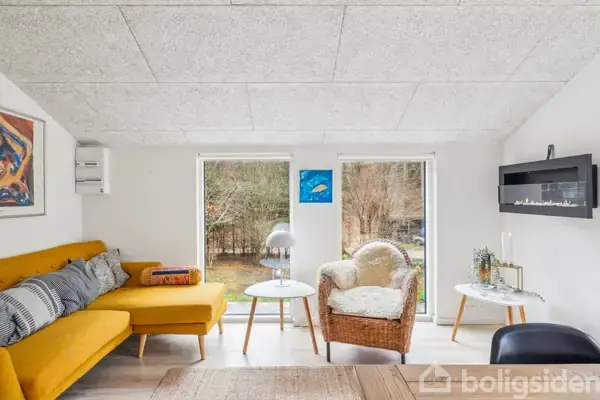 Gul sofa med blå og grå puder står i stue. Ved siden af, en kurvestol ved runde borde med lampe og dekorationer. Store vinduer åbner til haveudsigt. Kunst på væggene.