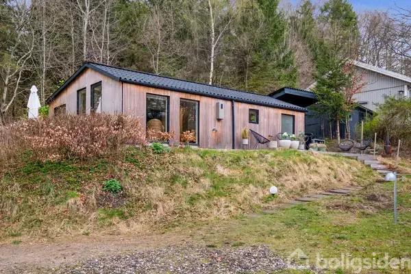 Et træhus står på en skråning, omgivet af græs og buske. Store vinduer vender mod en åben terrasse med havemøbler. I baggrunden ses en skovbevokset bakke.