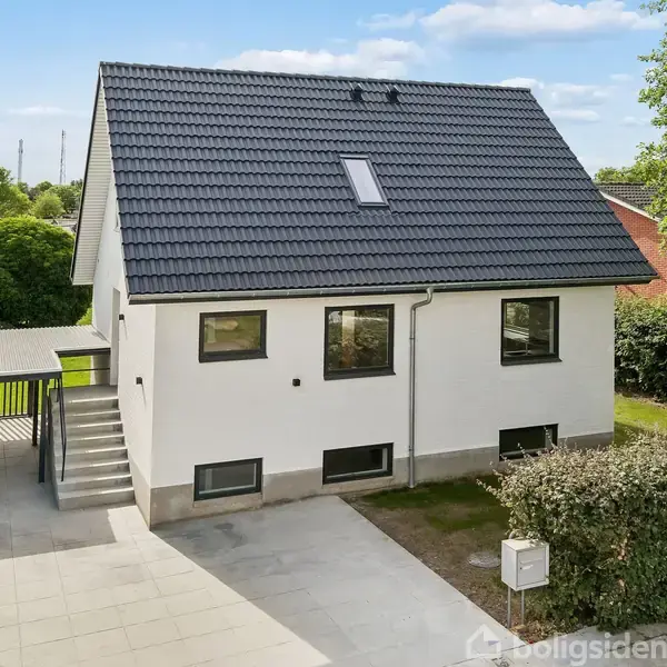 Et moderne, to-etagers hus med sort tag og hvid facade står i et beboelsesområde. Der er en indkørsel med fliser og en carport til venstre, omgivet af træer og hække.