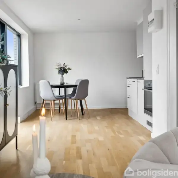 Et stilfuldt, lyst rum med et rundt bord og fire stole midt på et trægulv. Planten og lysestagerne tilføjer dekorative elementer. Køkkenfaciliteter ses til højre.
