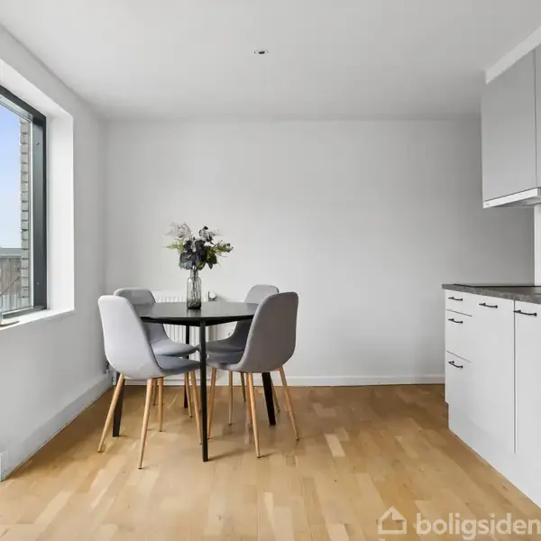 Et rundt spisebord med blomster i en vase står ved siden af fire stole. Bordet er placeret i et lyst køkken med trægulv og hvide køkkenskabe. Terrasse ses gennem det store vindue.