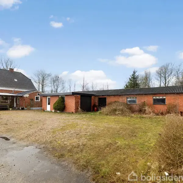 Hus med rødt murværk og sort tag står i en åben gårdsplads. Tilknyttede udhuse strækker sig til højre. Omgivet af træer og en græsplæne på en overskyet dag.