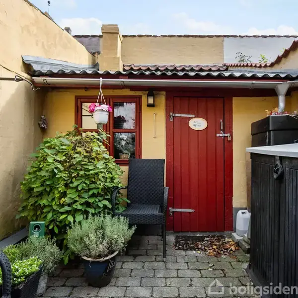 En rød dør med et lille vindue og skilt åbner til en gårdhave. To stole og blomsterkrukker står på stenfliser. Væggene er gule med et halvtag ovenfor.