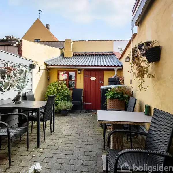 Terrasse med flisebelægning og sorte havemøbler placeret omkring borde. Omkransende gule mure og dekorative planter hængende på væggene. En rød dør fører til et andet område.