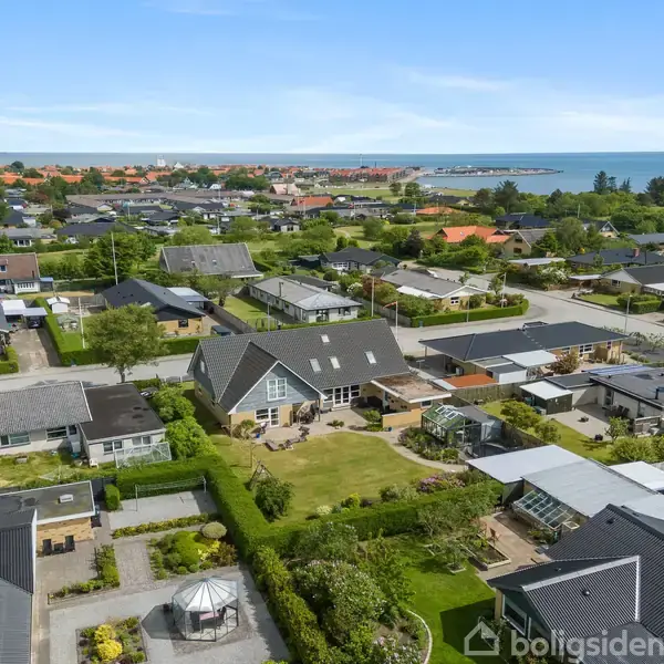 Flere huse strækker sig over en grøn boligområde, omgivet af haver og træer, med havet i horisonten og blå himmel ovenfor.