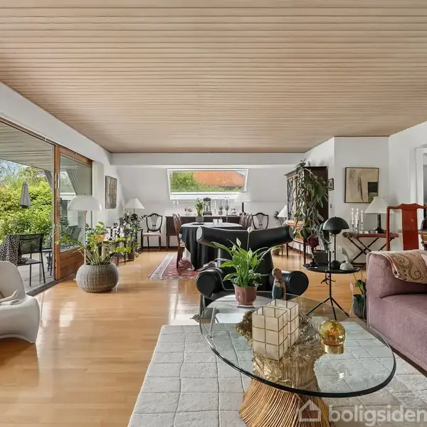 Stue med lilla sofa og glasbord med planter i forgrunden. Store vinduer giver udsigt til en græsplæne udenfor. Spisebord og stole i baggrunden skaber en åben og lys atmosfære.
