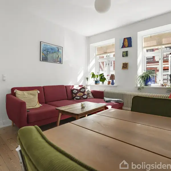 Rød sofa står i et lyst rum med trægulv, ved siden af et træbord med grønne stole. Sollys kommer ind gennem tre vinduer dekoreret med planter, plakater og malerier.