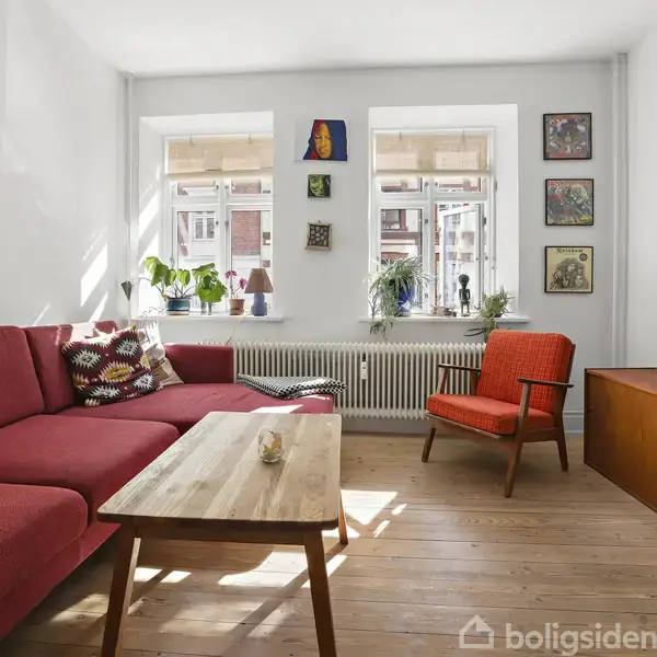 Rød hjørnesofa står i stue med trægulv, bøgetræsbord og rød lænestol. Vindue med grønne planter bagved, fladskærms-TV til højre. Væggene er pyntet med farverige malerier.