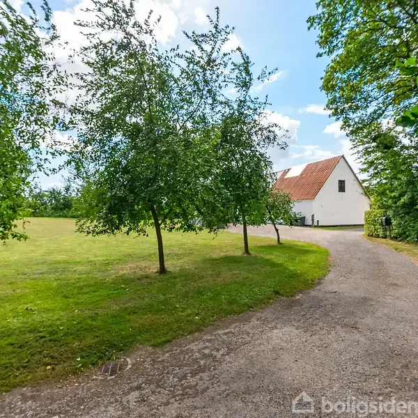 Hus med hvid facade og rødt tag står stille ved grusvej omgivet af grønne træer og græsplæne under en blå himmel.