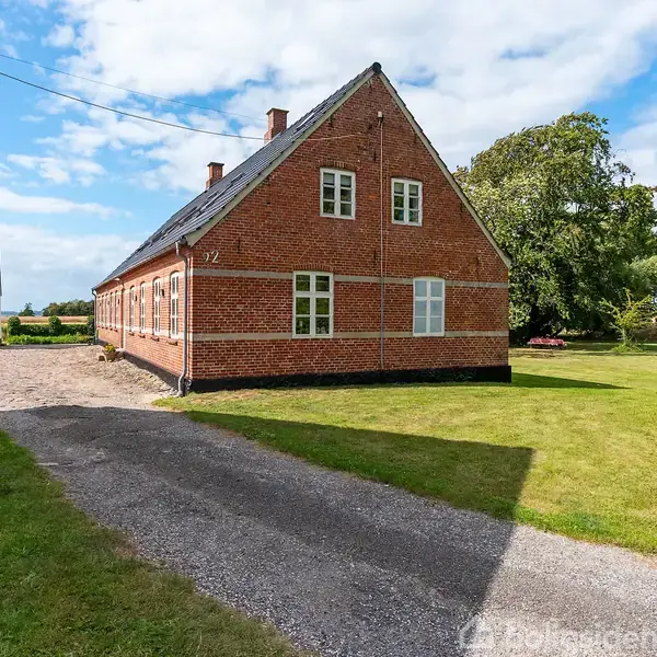 Rødt murstenshus med grønne vinduer og skråt tag står på en solrig, græsbevokset grund. Grusvej og anden bygning ses i baggrunden. Nummer "92" er synligt på huset.