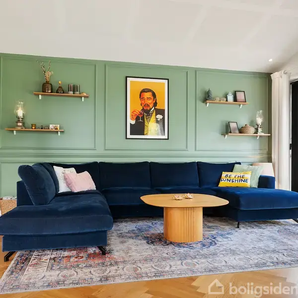 Mørkeblå sofa står mod grøn væg med hylder og kunst, inklusive et centralt portræt. Et rundt bord foran sofaen. Store vinduer med udsigt til grønne omgivelser. Pude med tekst: "BE THE SUNSHINE".