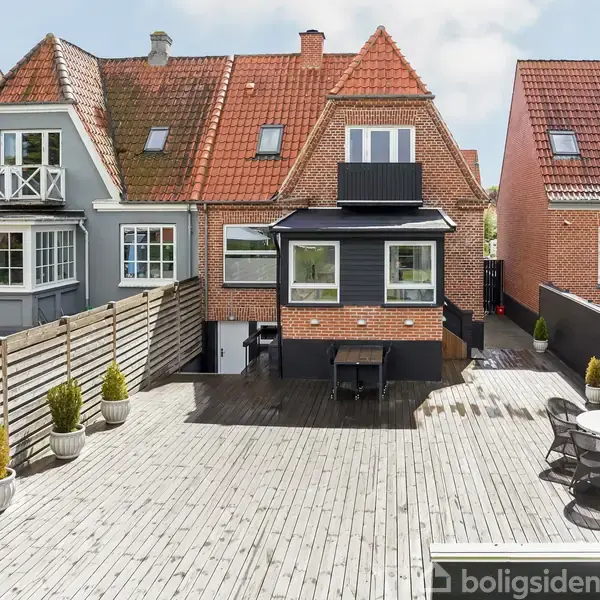To huse med røde tegltage står side om side, med en stor, træbelagt terrasse i forgrunden. Terrassen har borde, stole og planter langs et hegn.