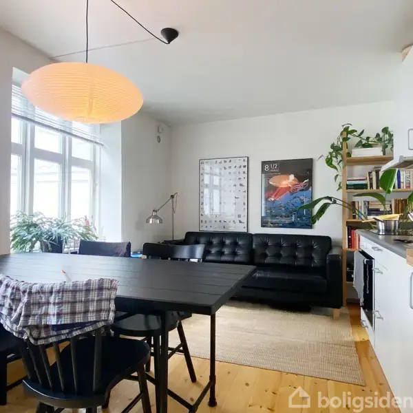 Et køkken-alrum med sorte møbler, en sort skinnesofa ved væggen, to plakater, et spisebord med stole og en loftlampe. Planter står ved vinduet; køkkenet til højre.