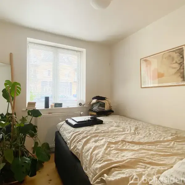 Soveværelset indeholder en dobbeltseng med en hvid dyne, et spejl og en stor plante ved siden af et vindue med persienner. Der er en ramme på væggen.