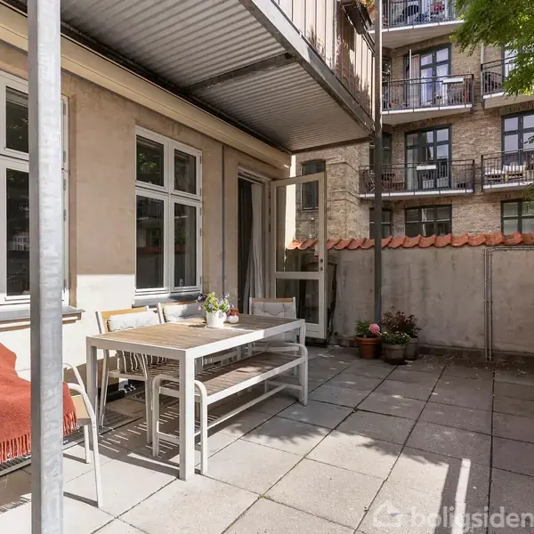 Terrasse med et bord og stole, placeret foran store vinduer. Et tæppe ligger over en stol. Omkring er der en murstensbygning og planter i krukker ved væggen.