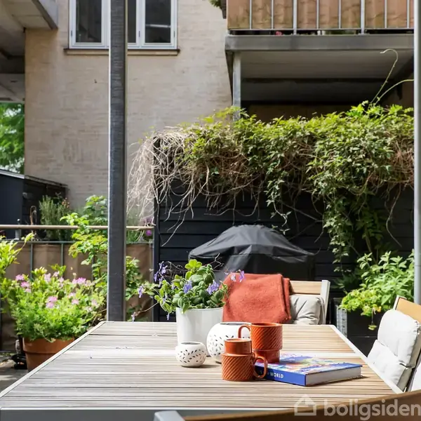 Et terrasseområde med et bord fyldt med en bog, kopper og en plante i en beholder. Omkring bordet er der stole og frodige planter, med en bygning i baggrunden.