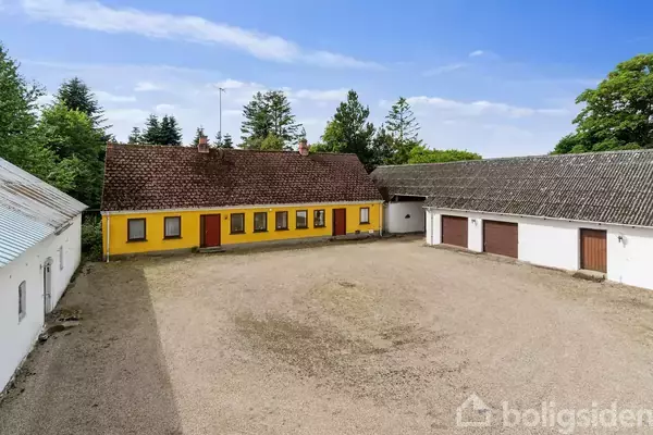 En gul murstensbygning og hvide lader står rundt en gårdsplads med grus. Træer omgiver stedet, der ser landligt og stille ud.