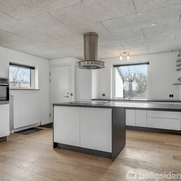 Et moderne køkken med en stor, central køkkenø i hvidt og sort. Rummet har trægulv, indbygget ovn, to vinduer og en emhætte over køkkenøen. Trehyldet væghylde til højre.
